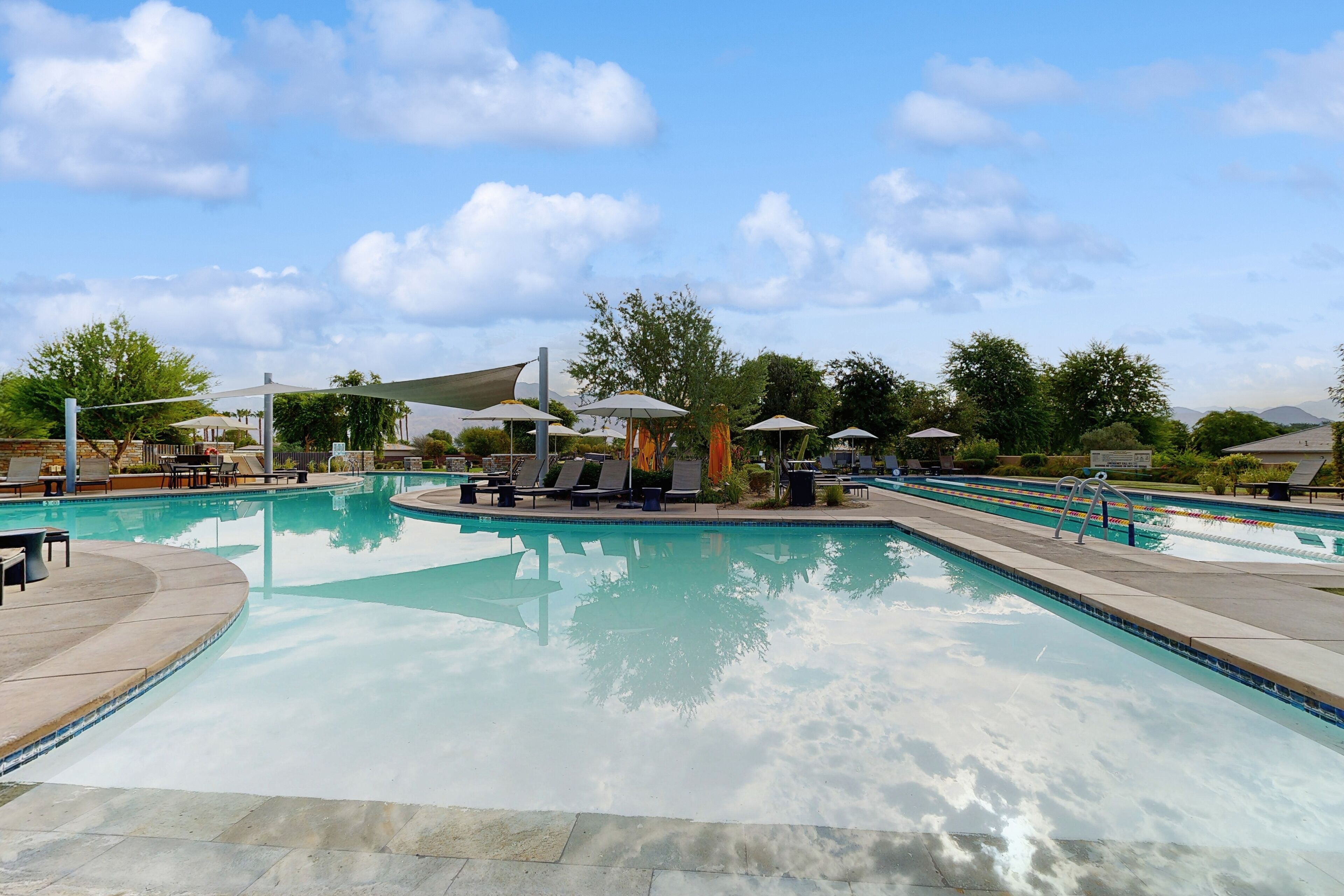 A swimming pool area with lounge chairs and umbrellas, surrounded by trees and a clear sky.