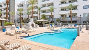 Una piscina con tumbonas y palmeras, rodeada por un edificio de varios pisos con balcones.