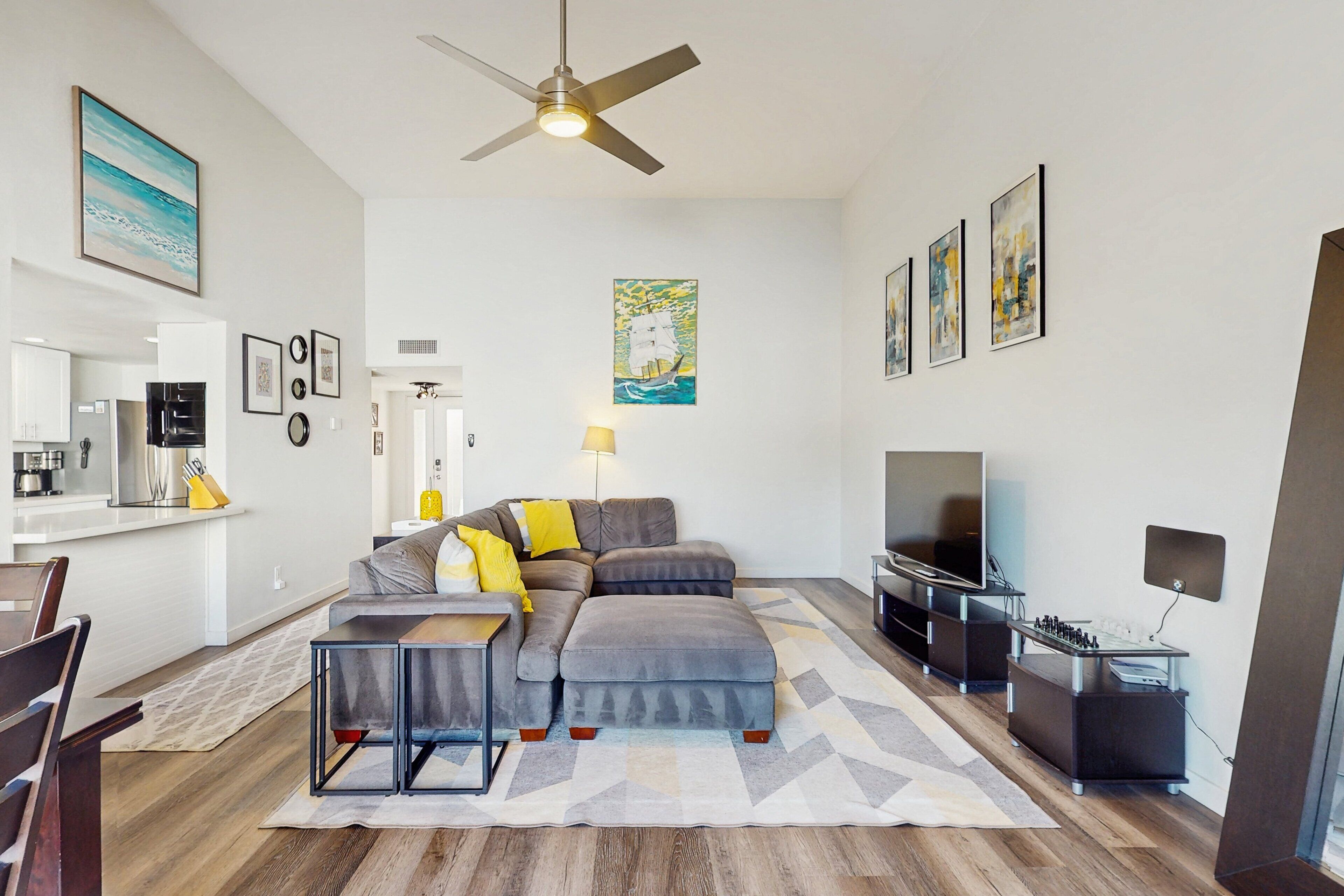 Un salon moderne avec un canapé d’angle, un téléviseur à écran plat, un ventilateur de plafond et des œuvres d’art accrochées aux murs.