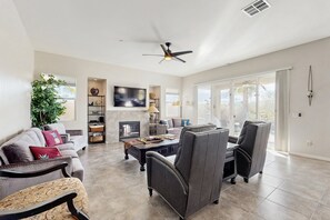 A spacious living room with a fireplace, comfortable seating, a flat-screen TV, and a large window offering a view. - Mountain view home, private pool & spa - shared pools, tennis, fitness center (Cathedral City)