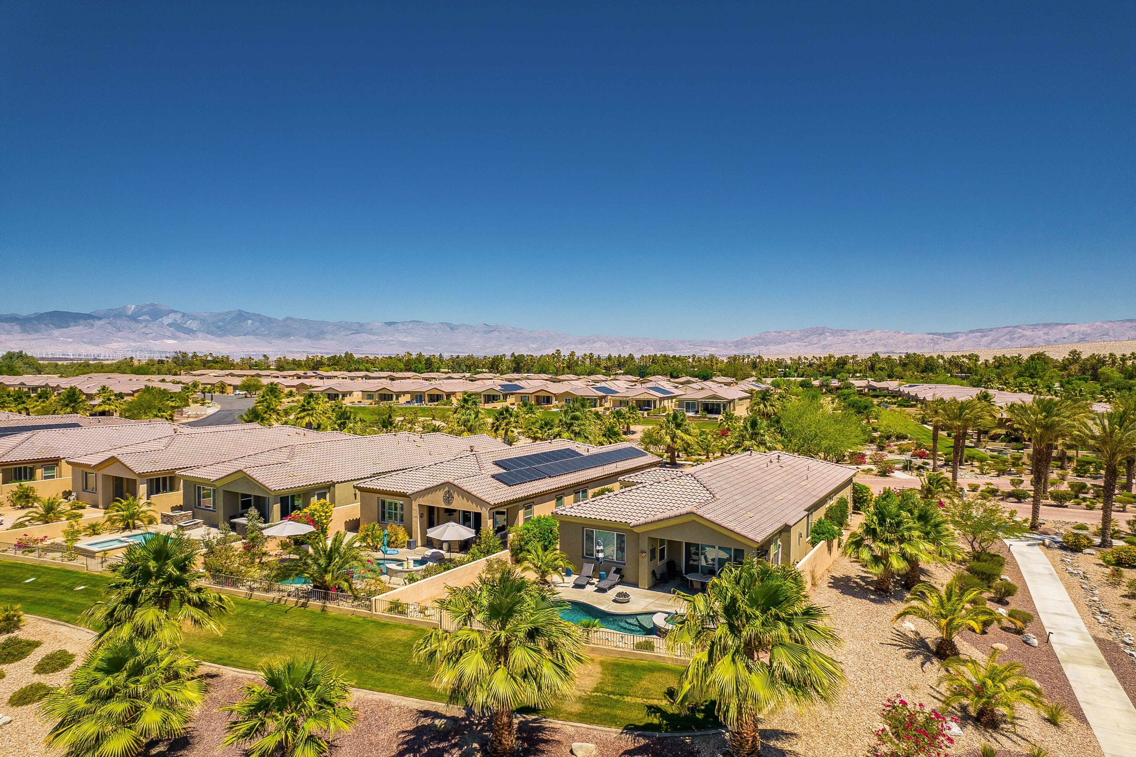 A resort with multiple villas, each featuring a swimming pool, surrounded by palm trees and a clear view of distant mountains.