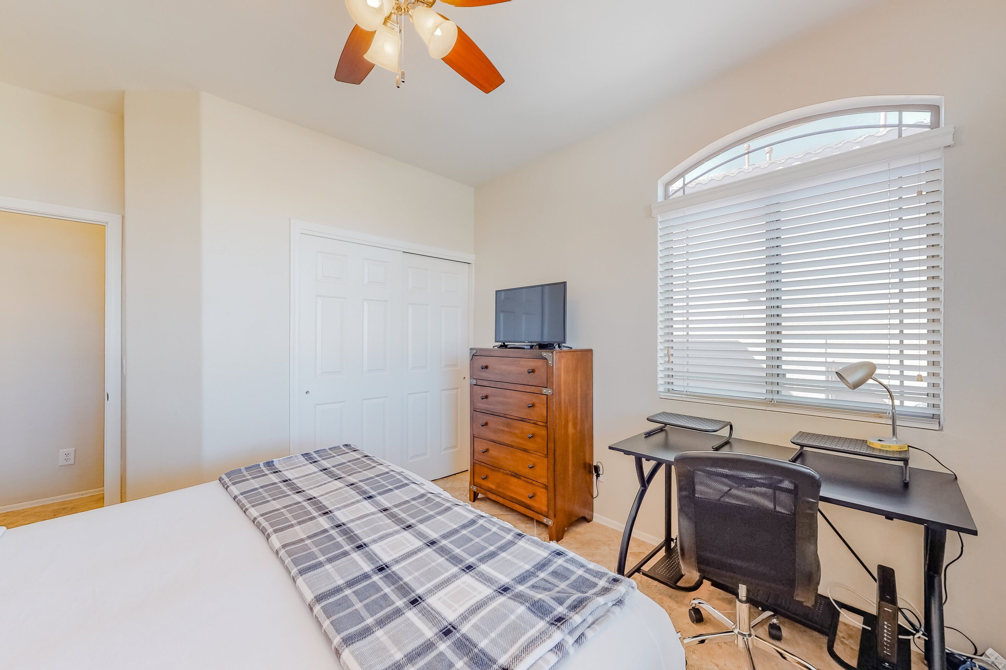 A bedroom with a bed, a desk with a chair, a dresser, a ceiling fan, and a window with blinds.