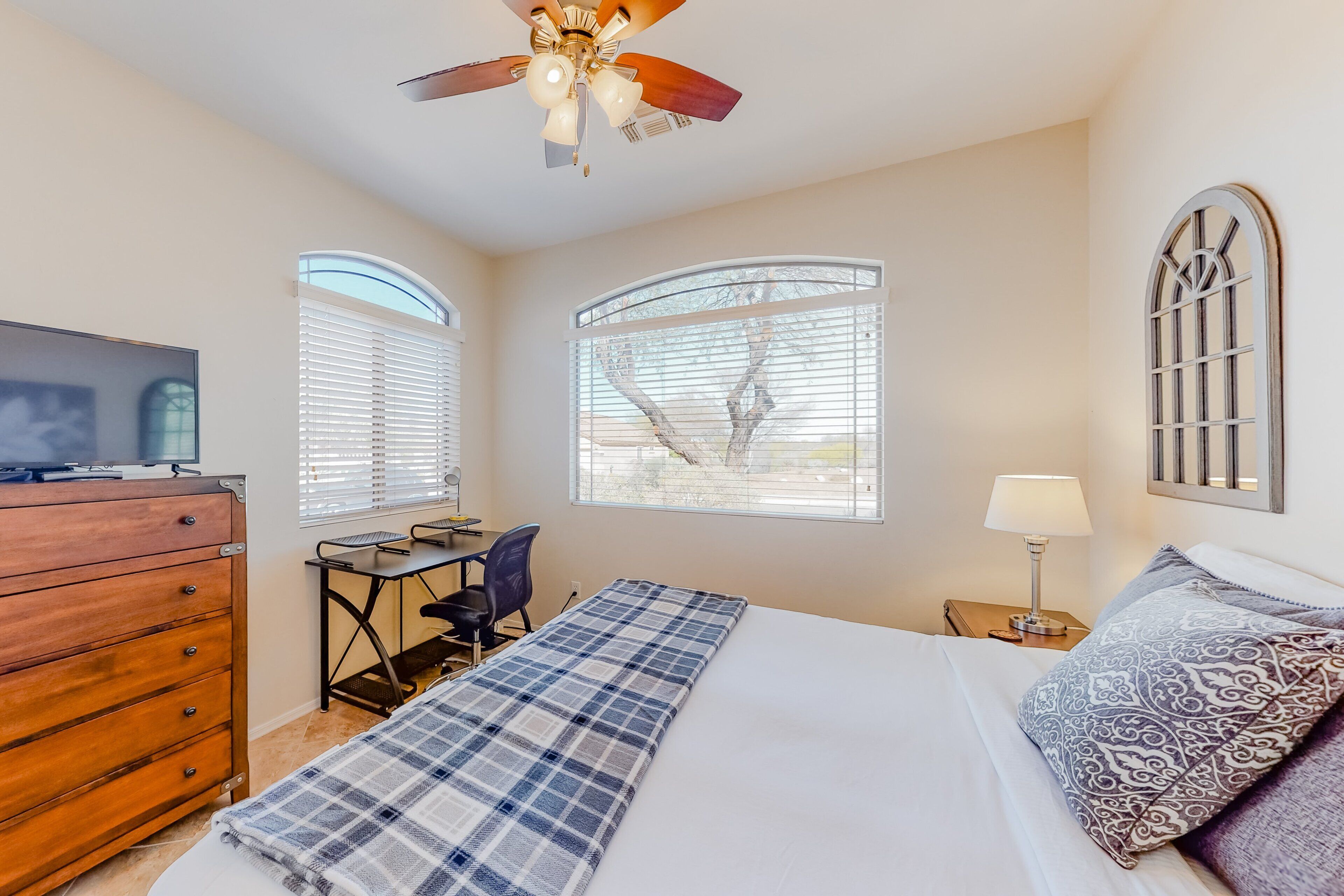 A bedroom with a bed, a desk, a chair, a dresser, a ceiling fan, and two windows.