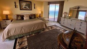 Quarto de hotel com cama, penteadeira, cadeira, ventoinha no teto, janela grande com vista e quadro na parede.