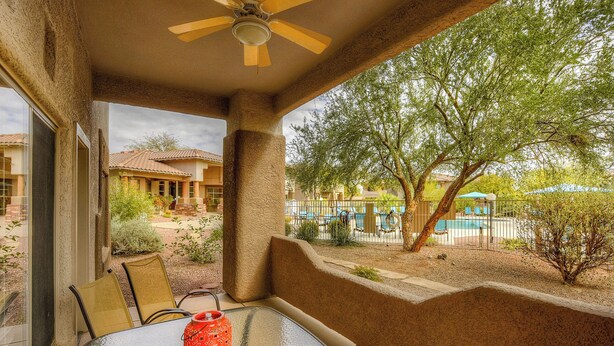 A covered patio with a table and chairs, a ceiling fan, and a view of a swimming pool and trees. - Comfortable condo with renovated kitchen, patio, pool, hot tub & fitness center (Oro Valley)