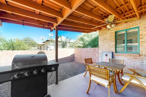 An outdoor patio with a grill, wooden table, and chairs, a brick wall, and a window with green trim. - Stylish Sam Hughes Escape With Private Yard, Dogs OK & Free WiFi (Tucson)