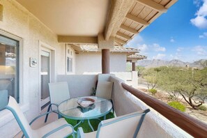 A balcony with a glass table and chairs, a view of the desert landscape, and a building with a stucco exterior. - Renovated condo with updated floors, pool, hot tub & mountain views (Oro Valley)