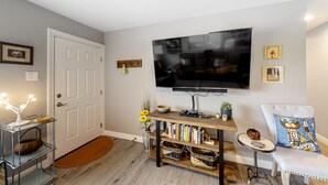 A living room with a flat-screen TV mounted on the wall, a bookshelf, a small side table, a chair with a decorative pillow, and a door with a doormat. - Snowbird-friendly home with central AC, washer/dryer & free WiFi - near golf (Phoenix)