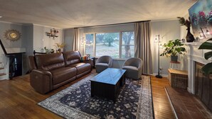 Ein Wohnzimmer mit braunem Ledersofa, einem Couchtisch, zwei Sesseln, einem gemusterten Teppich, einem Kamin und einem großen Fenster mit Vorhängen.