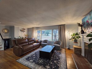 A living room with a brown leather sofa, a coffee table, two armchairs, a patterned rug, a fireplace, and a large window with curtains. - Great location: UT, Windorck, national lab, melton lake park (Oak Ridge)