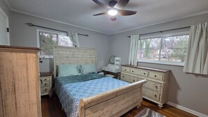 Ein Schlafzimmer mit einem hölzernen Bett, einem Schminktisch, einem Nachttisch, einem Deckenventilator und einem Fenster mit Vorhängen.