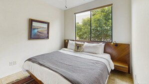 Une chambre à coucher avec un lit, un ventilateur de plafond, une fenêtre donnant sur des arbres et un tableau encadré au mur.