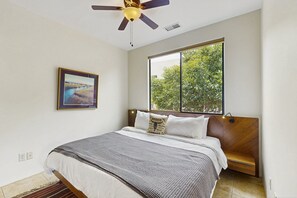Ein Schlafzimmer mit Bett, Deckenventilator, Fenster mit Blick auf Bäume und einem gerahmten Bild an der Wand.