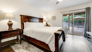 A bedroom with a wooden bed, two bedside tables, a ceiling fan, and a view through a glass door to an outdoor area with umbrellas. - Dog-friendly home with enclosed yard, pool, putting green, grill, foosball, & AC (Scottsdale)
