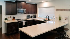 A modern kitchen with dark wood cabinets, a stainless steel microwave, a large island with stools, and a 'Gather Together' sign on the wall. - Dog-friendly home with enclosed yard, pool, putting green, grill, foosball, & AC (Scottsdale)