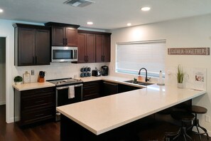 A modern kitchen with dark wood cabinets, a stainless steel microwave, a large island with stools, and a 'Gather Together' sign on the wall. - Dog-friendly home with enclosed yard, pool, putting green, grill, foosball, & AC (Scottsdale)