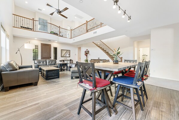 A spacious living area with a high ceiling, a dining table surrounded by chairs, a staircase to an upper level, and a modern kitchen in the background.