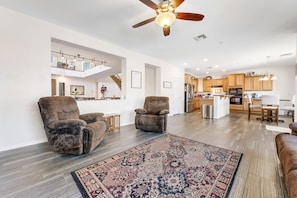 A spacious living room with two plush armchairs, a patterned rug, and a ceiling fan. A kitchen and dining area are visible in the background.