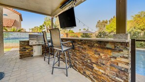 Un patio exterior con una barbacoa de piedra, dos taburetes de bar, un televisor de pantalla plana montado en la pared y una piscina al fondo.