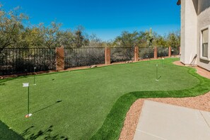 A well-maintained artificial grass golf course with a clear sky, a cactus in the background, and a building on the right.