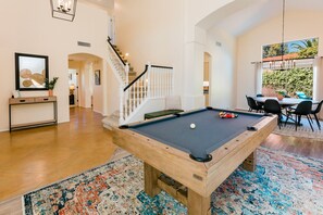A pool table in a spacious room with a wooden floor, a staircase, and a dining area with chairs and a table.