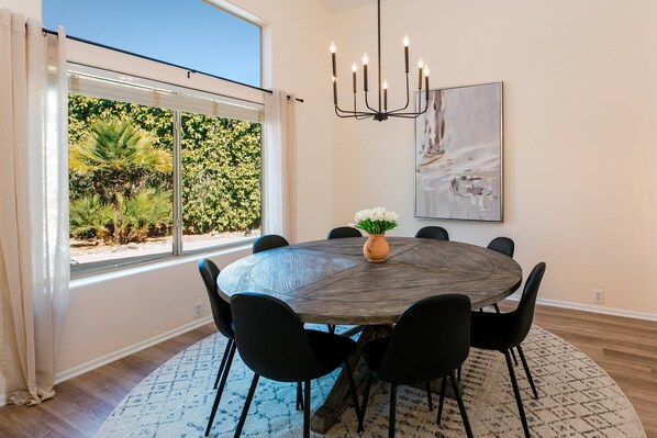 A dining area with a round table, black chairs, a chandelier, a potted plant, and a window with a view of greenery.