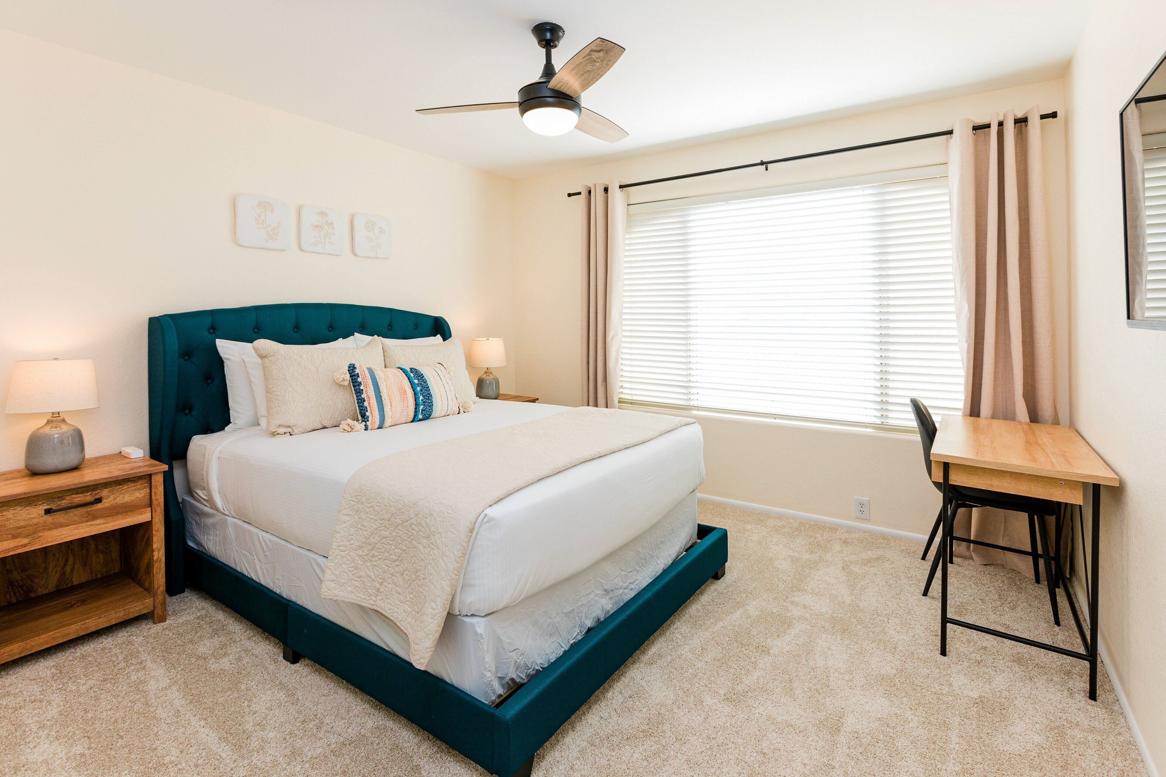 A bedroom with a bed, bedside table, lamp, ceiling fan, window with curtains, and a desk with chair.