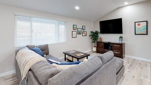 A modern living room with a grey sofa, a coffee table, a TV mounted on the wall, and a window with blinds. - Modern Ranch with Private Putting Green, Fast WiFi, Central AC, & Great Location (Scottsdale)