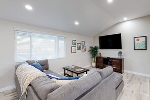 A modern living room with a grey sofa, a coffee table, a TV mounted on the wall, and a window with blinds. - Modern Ranch with Private Putting Green, Fast WiFi, Central AC, & Great Location (Scottsdale)