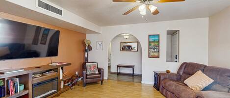 A living room with a television, leather sofa, wooden flooring, ceiling fan, and a fireplace.