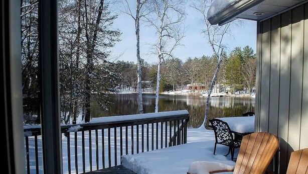 A wooden deck with snow-covered chairs and a table, overlooking a calm lake and trees. - Mapleview - Three Bedroom Cottage, Sleeps 12 (Cloyne)