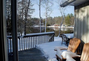 A wooden deck with snow-covered chairs and a table, overlooking a calm lake and trees.