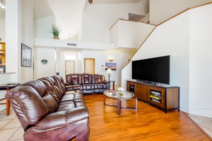 Ein geräumiges Wohnzimmer mit einem braunen Ledersofa, einem Glastisch und einem Flachbildfernseher auf einer Holzkommode.