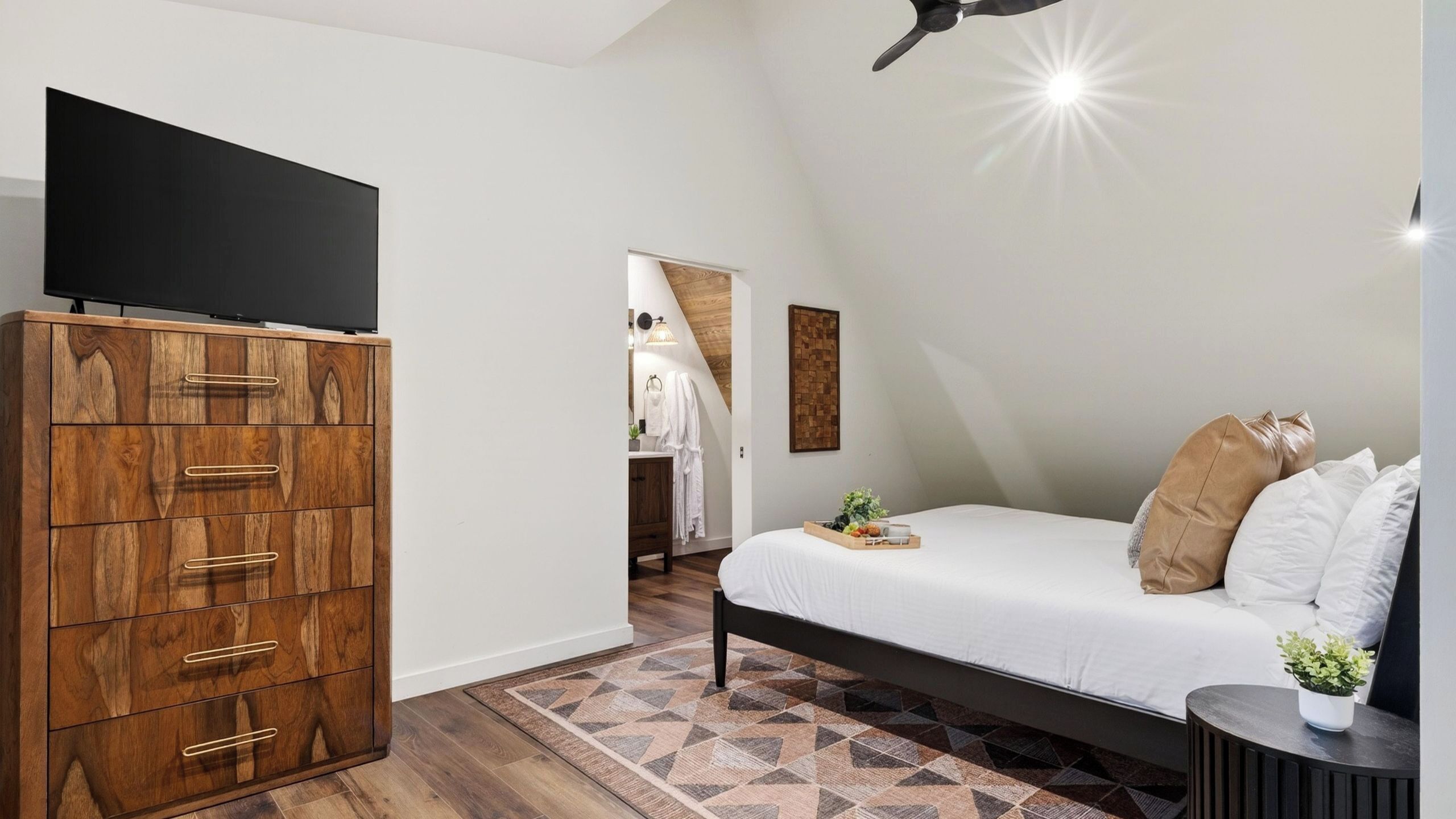 A bedroom with a bed, a wooden wardrobe, a television, a ceiling fan, and a geometric patterned rug.