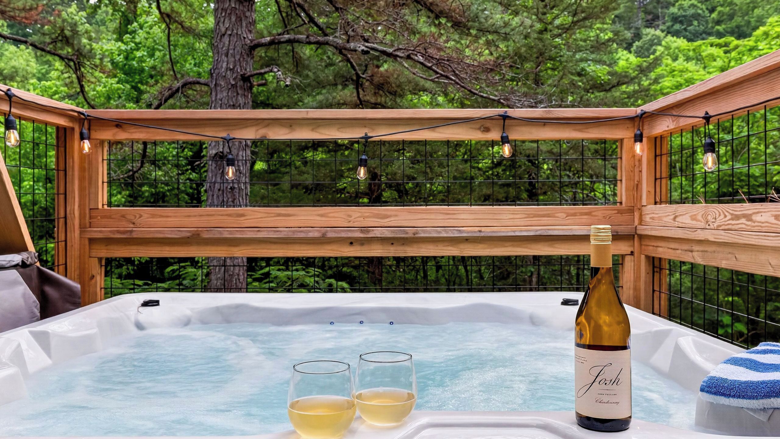 A hot tub with two glasses of white wine and a bottle, surrounded by a wooden deck with string lights, set against a backdrop of trees.