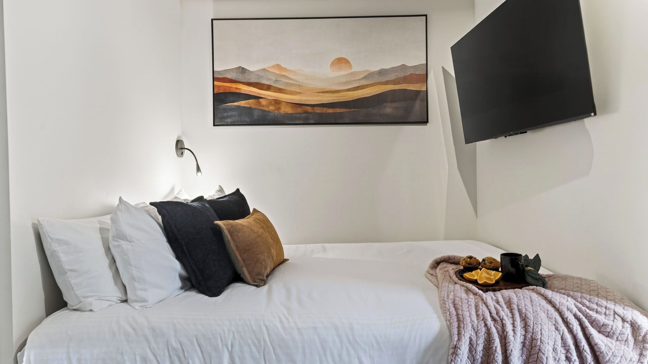 A bedroom with a bed, pillows, a blanket, a painting, and a TV mounted on the wall.