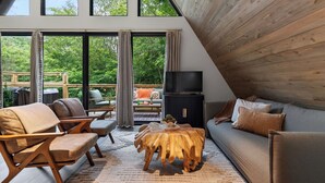 Ein gemütliches Wohnzimmer mit einer Couch, Sesseln, einem Holztisch und einem Fernseher. Große Fenster bieten einen Blick ins Freie.