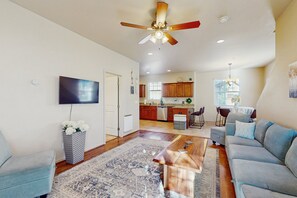 Ein geräumiges Wohnzimmer mit Deckenventilator, Flachbildfernseher, einer Couch, einem Couchtisch und einer Küche im Hintergrund.