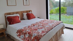 Quarto com cama, almofadas, mesa de cabeceira, luminária e janela com vista para o verde.