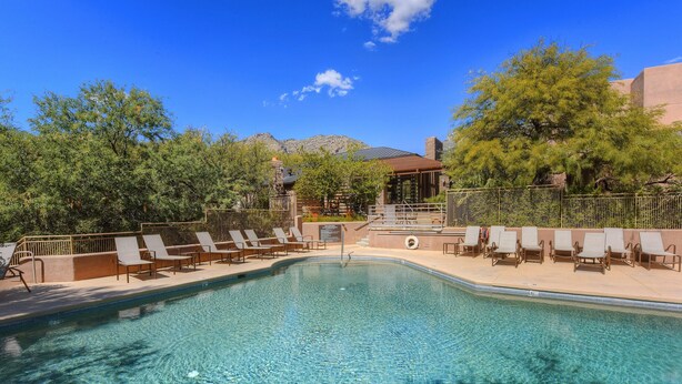 A clear swimming pool with lounge chairs and a wooden deck, surrounded by trees and a building in the background.