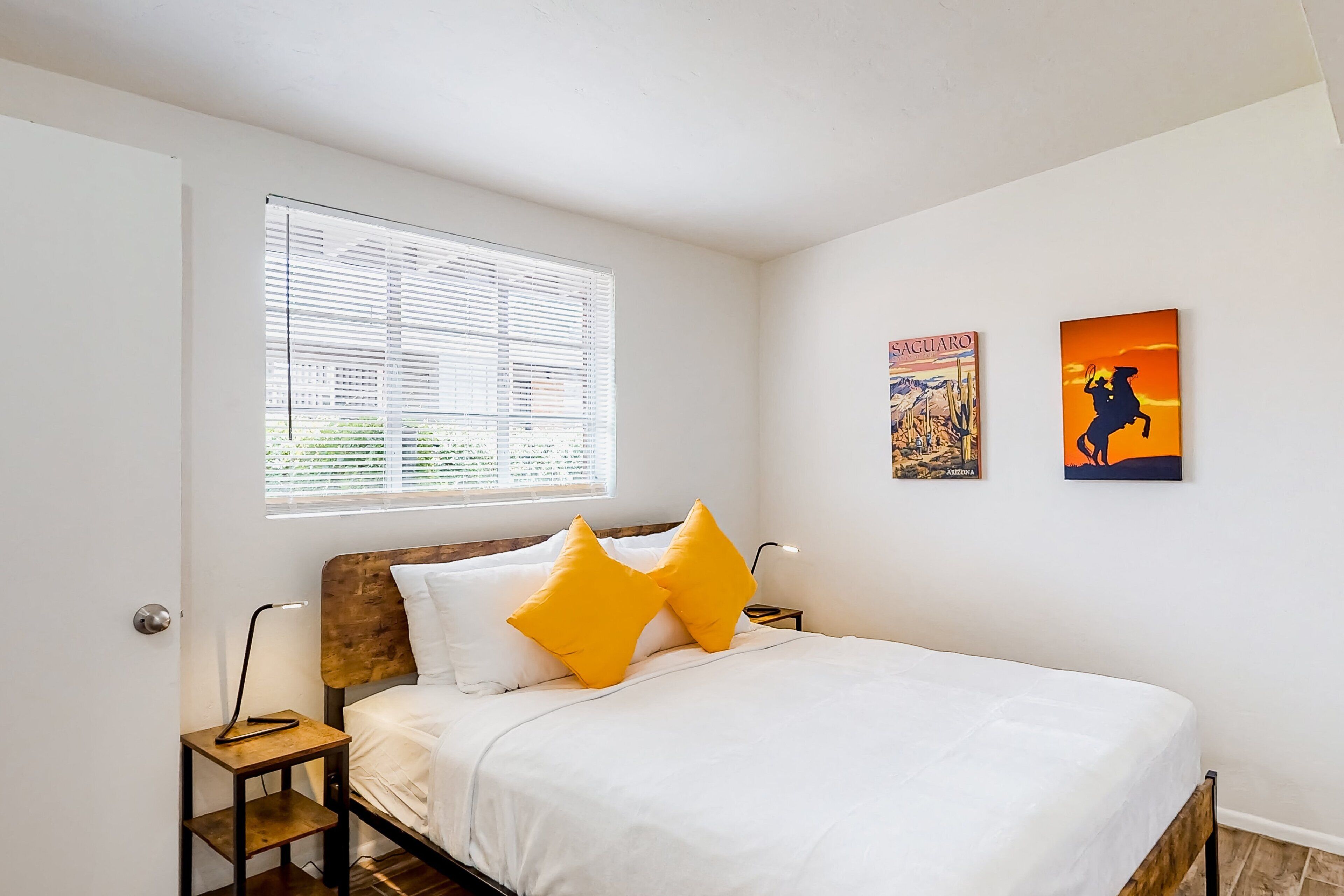 Un dormitorio con cama, mesita de noche, lámpara, ventana con persianas y dos cuadros en la pared.