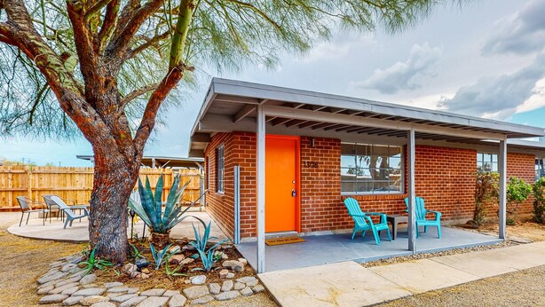 Ein modernes Haus mit Backsteinfassade, einer überdachten Terrasse mit blauen Stühlen und einem Baum mit Agavenpflanze.
