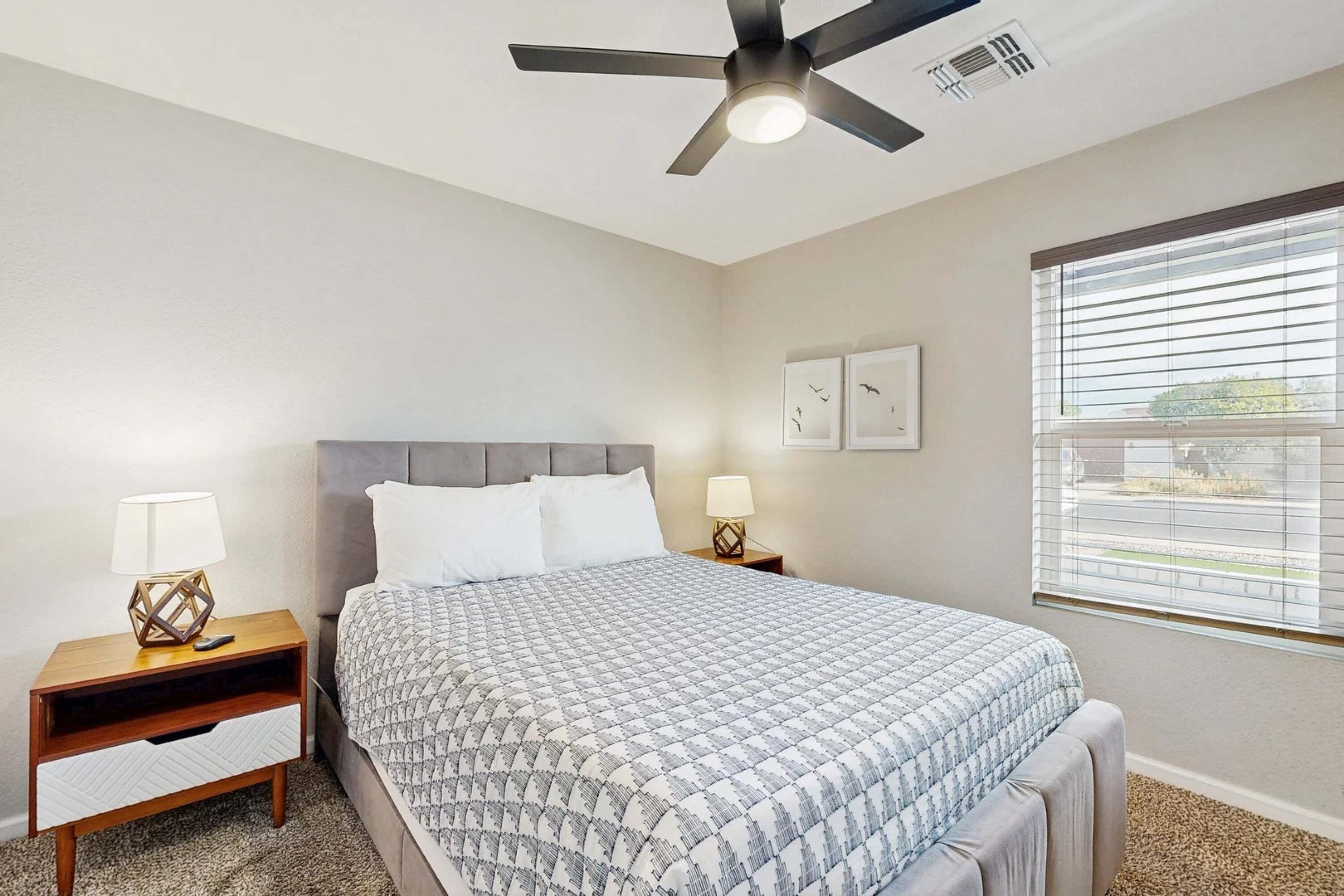A bedroom with a bed, two bedside tables with lamps, a ceiling fan, a window with blinds, and wall art.