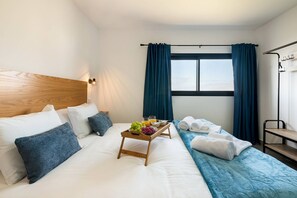 Een hotelkamer met een bed, een houten hoofdbord, een nachtkastje met een schaal fruit en een glas sinaasappelsap, een blauwe spreidek en een raam met gordijnen.