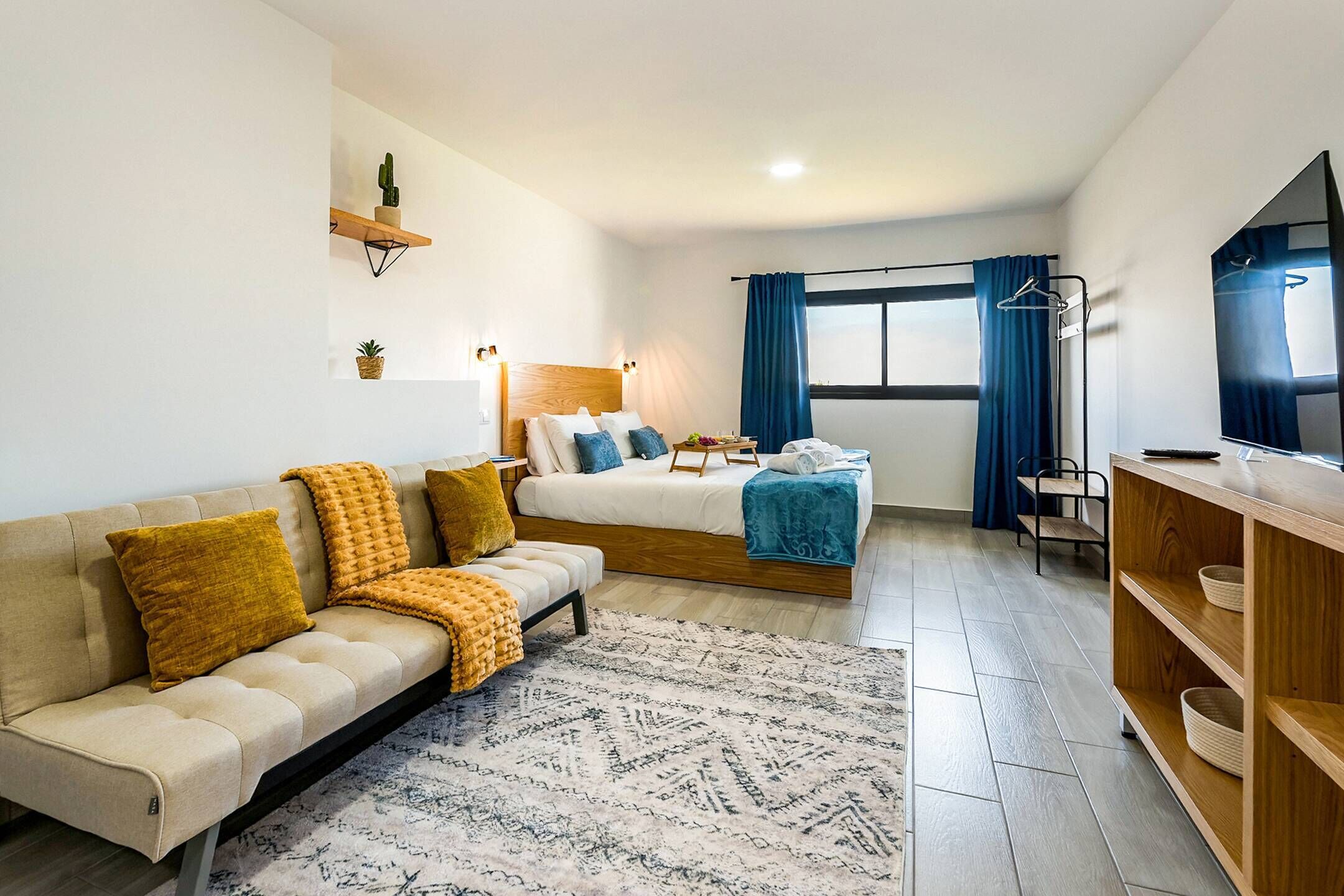 A modern hotel room with a sofa, a bed with a wooden headboard, a TV, and a wooden shelf.