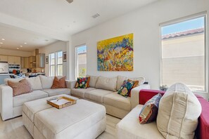 A spacious living room with a sectional sofa, a red armchair, and a coffee table. A large abstract painting adorns the wall, and the room has large windows offering a view of the outside. - Bright desert oasis  w/hot tub & grill - shared pool & tennis in  55+ community (Rancho Mirage)