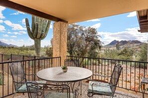 A patio with a round table and chairs, a cactus, and a view of mountains and desert vegetation.
