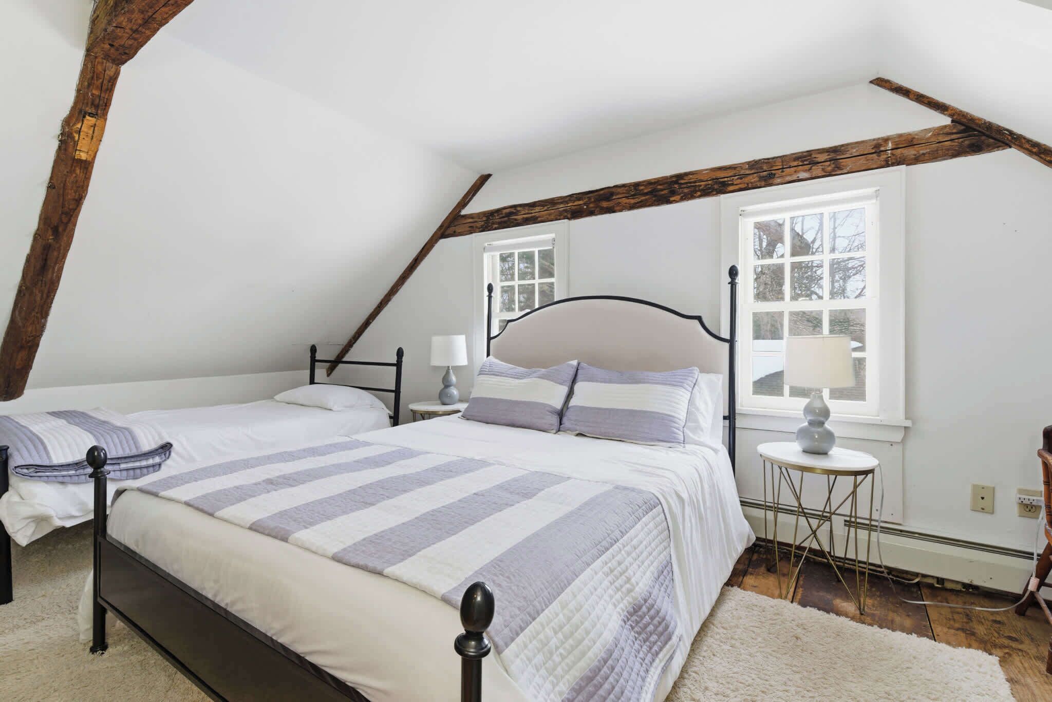 A bedroom with two beds, a wooden ceiling beam, a window, and a small table with a lamp.