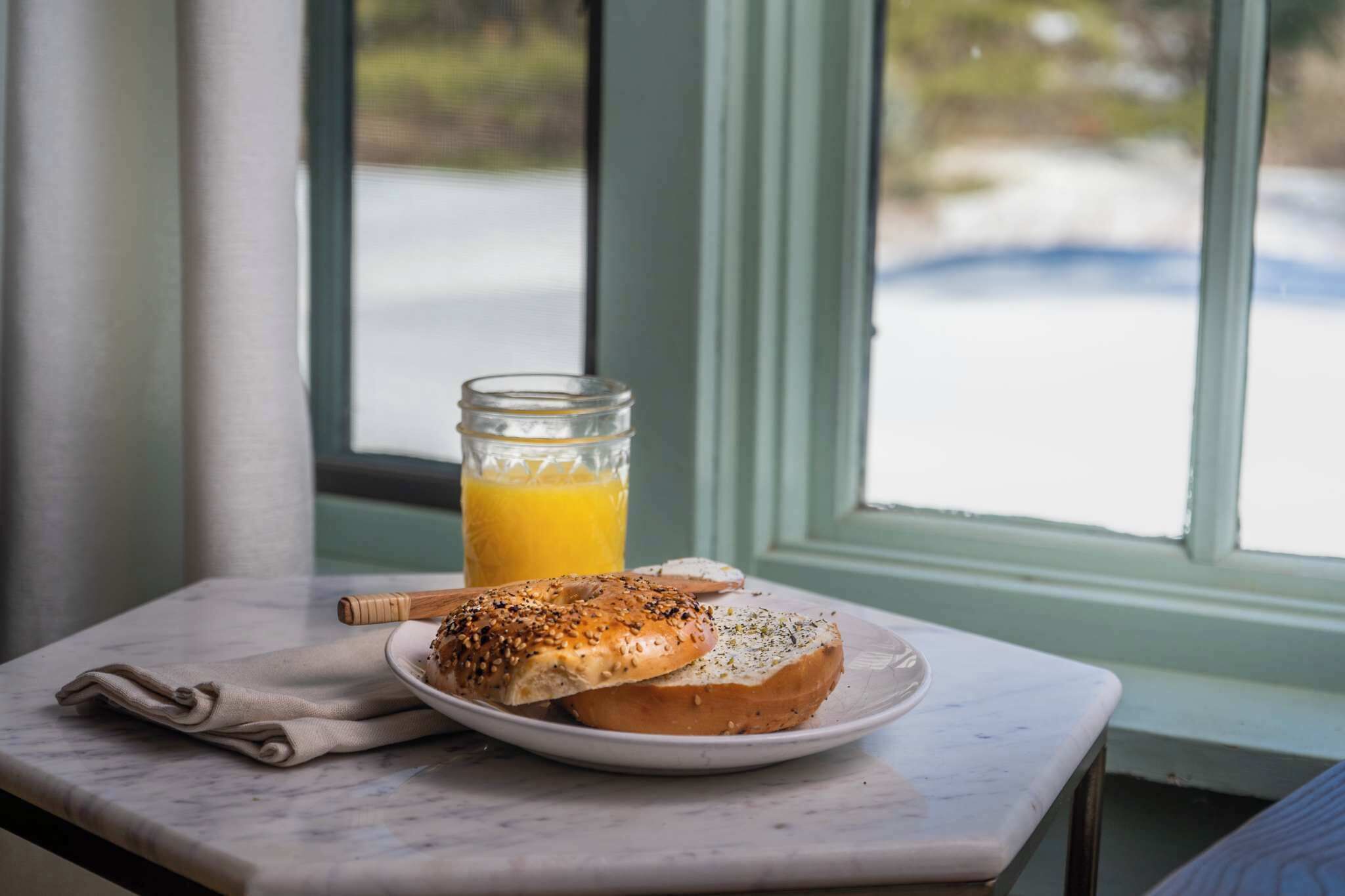 Un desayuno con bagels en un plato, un vaso de jugo de naranja y una rama de canela sobre una mesa de mármol cerca de una ventana.