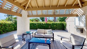 En udendørs terrasse med et træpergola, en sofa, to lænestole, et bord og et flag.